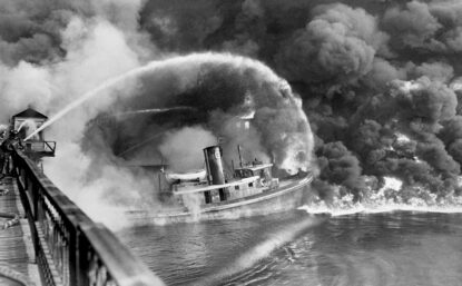 Black and white photograph of a boat on fire in a river and a small hose attempting to extinguish it.