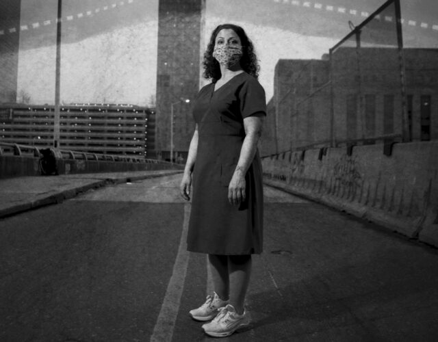 Black and white photograph of nurse Linda Ruggiero wearing a face mask, standing outside on a bridge.